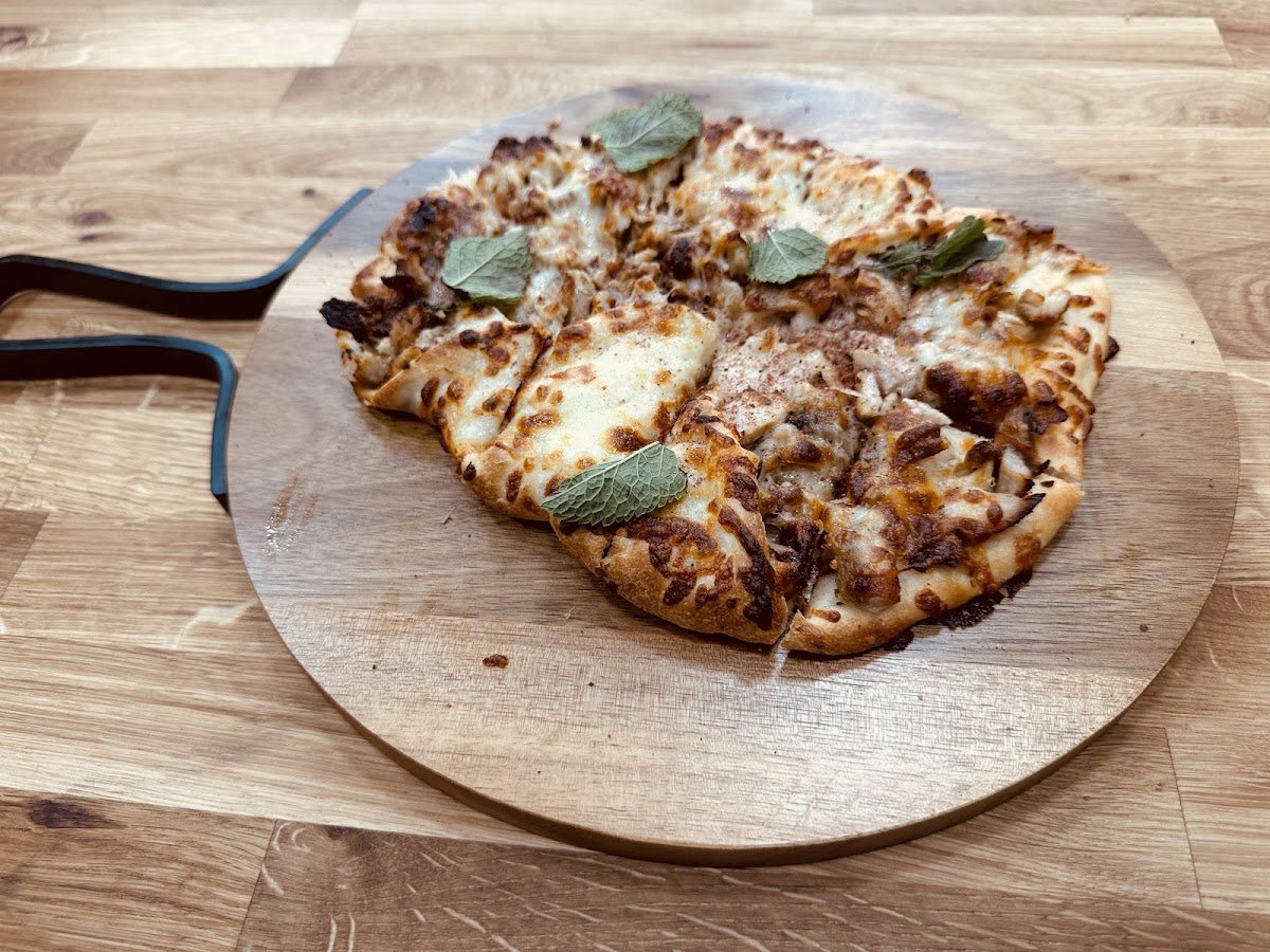 A golden, cheese-topped flatbread cut into slices and scattered with fresh leaves, on a round wooden board.