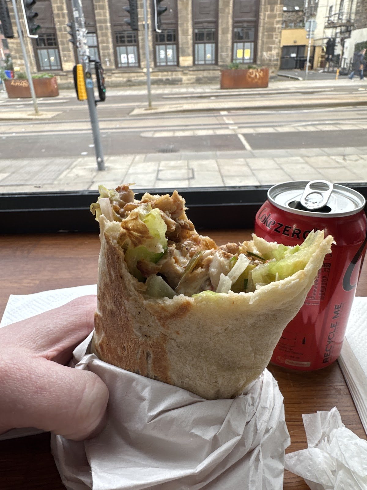 A shawarma wrap bitten open, stuffed with meat, lettuce and sauce, held at the window counter with Leith Walk and its tram stop outside.