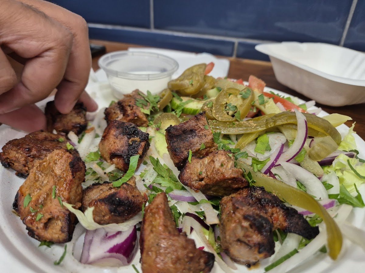 Charred grilled meat pieces on a bed of salad with sliced red onion, parsley and pickled green chillies, with a pot of white sauce.